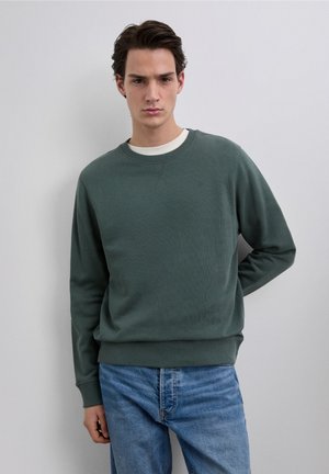 Groene trui met geribbelde ronde hals, gestructureerde stof en lange mouwen, gecombineerd met een blauwe spijkerbroek. Minimalistisch ontwerp met subtiele branding.