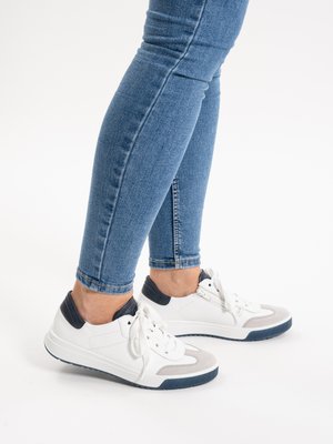 Legs in blue skinny jeans wearing white sneakers with navy and gray accents, standing on a white background.
