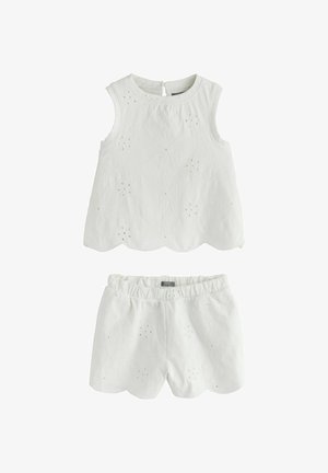 Fată tânără cu păr lung și închis la culoare poartă un top alb fără mâneci și pantaloni scurți cu broderie florală, stând pe un fundal bej.