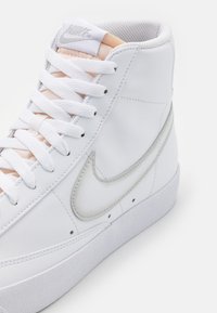 Bílé vysoké tenisky vyrobené z hladké kůže, s stříbrným logem Nike, bílými tkaničkami a texturovanou gumovou podrážkou.