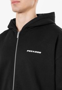 Crna zip-up dukserica od pamuka, s prednjim srebrnim patent-zatvaračem i bijelim logotipom "PEGADOR" na prsima. Jednostavan dizajn, bez uzoraka.