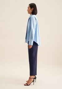 Camicia oversize a righe blu e bianche, maniche lunghe, abbinata a pantaloni scuri navy a gamba affusolata e sandali con tacco neri. Tessuto con texture liscia.