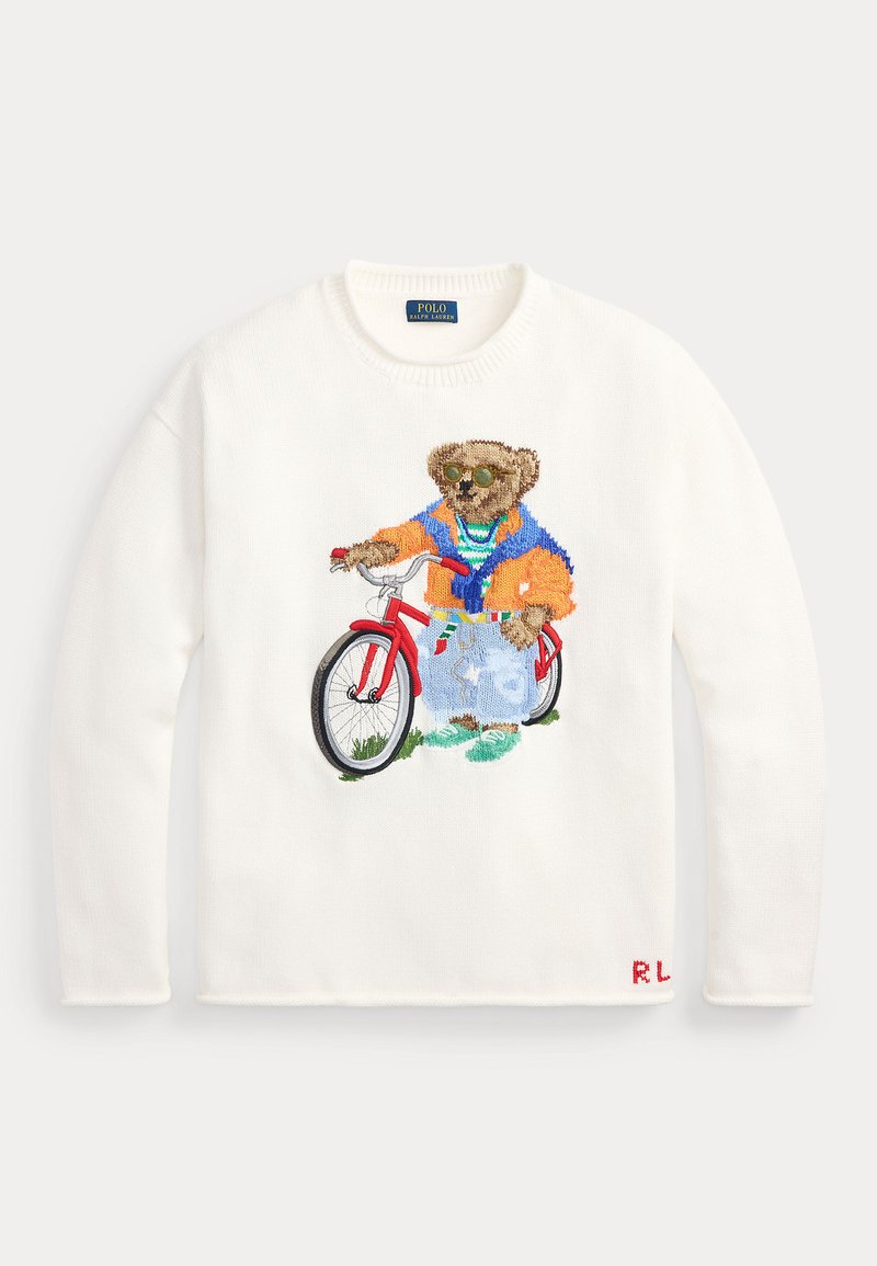 Pull blanc avec un ours brodé portant des lunettes de soleil, une veste colorée et un jean, debout à côté d'un vélo rouge.