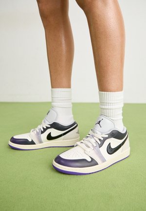 Jalat, joissa valkoiset polven alapuoliset sukat ja mustavalkoiset sekä violetit Nike Air Jordan -lenkkarit seisomassa vihreällä lattialla.