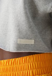 Gros plan sur une personne portant un sweat-shirt gris raccourci avec l'inscription « BORG » et un short à taille smockée orange vif.