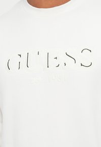 Acercamiento de una camisa blanca con el logotipo "GUESS EST. 1981" en relieve y bordado en el pecho.