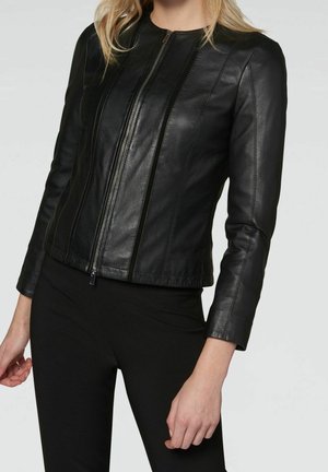 Femme portant une veste en cuir noire cintrée avec une fermeture éclair sur le devant, associée à un pantalon noir, sur un fond clair uni.