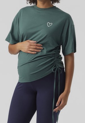 Gravid person iført en grøn rynket sidetshirt med et lille hjerte-logo, hvilende den ene hånd på maven og den anden ved siden af kroppen.
