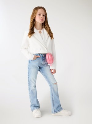 Ragazza giovane con capelli lunghi che indossa un blazer corto bianco, jeans chiari strappati, sneakers bianche e una piccola borsa a tracolla rosa.
