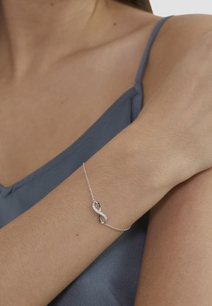 Silbernes Armband mit einem Unendlichkeitssymbol, das mit eingearbeiteten Strasssteinen verziert ist. Feine Kette liegt auf einem nackten Handgelenk und wird durch ein hellblaues Oberteil ergänzt.