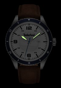 Reloj Timberland con esfera plateada, números en negro y bisel azul. Correa de cuero marrón. Manos y logotipo luminiscentes en la esfera.