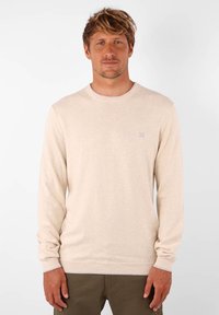 Pull beige clair en tricot doux, avec un col rond et des manches longues, arborant un logo discret sur la poitrine.