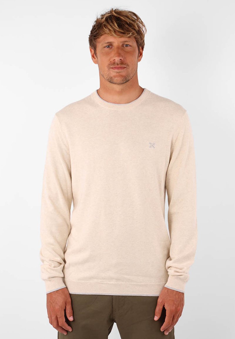 Pull beige clair en tricot doux, avec un col rond et des manches longues, arborant un logo discret sur la poitrine.