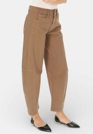 Brązowe, wysokie spodnie zwężane z przodu i po bokach z kieszeniami, noszone z czarnymi balerinami z ostrym noskiem na jasnym tle.