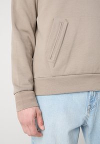 Haut beige texturé à manches longues avec détail de poche zippée, associé à un jean denim bleu clair, montrant une partie de la main et du bas du torse.