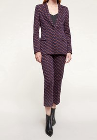 Marinblå kostym med orange geometrisk design; skräddarsydd kavaj med en knapp och slagkragar; skräddarsydd kort byxa; svarta ankelstövlar.