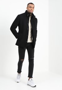 Schwarzer Wollmischungenmantel mit Knopfverschluss, kombiniert mit einem cremefarbenen Pullover und zerrissenen schwarzen Jeans. Das Model trägt weiße Sportschuhe.