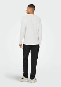 Weißes Langarmhemd mit strukturiertem Muster, kombiniert mit schwarzen Hosen, getragen mit cremefarbenen Sneakers, vor einem hellen Hintergrund stehend.