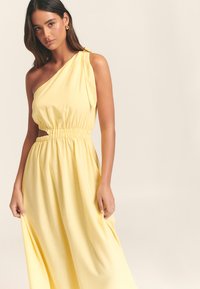 Keltainen yksialkainen mekko, jossa on smokkailtu vyötärö ja leikkausdetaili. Valmistettu pehmeästä, laskeutuvasta kankaasta, jossa on lempeä kiilto ja laskeutuvuus.