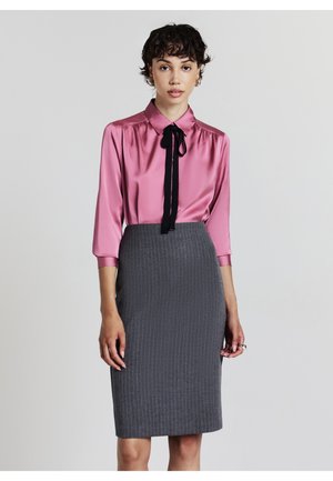 Blouse en satin rose avec une cravate noire, associée à une jupe crayon à rayures grises. La blouse a un col et des manches trois-quarts.
