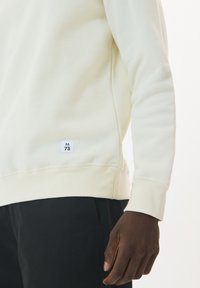 Lichtgekleurde sweatshirt gemaakt van zacht materiaal, met een aansluitend ontwerp, geribde mouwen en een klein label met "M 73" aan de onderkant.