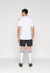 Λευκό αθλητικό πουκάμισο με κόκκινες λεπτομέρειες, μαύρο σορτς και λευκές κάλτσες μέχρι το γόνατο με μαύρες ρίγες. Παρουσιάζεται από πίσω.