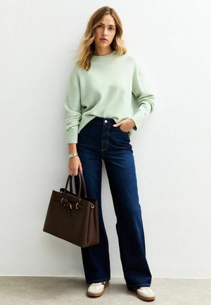 Muntgroene oversized trui, hoog-getailleerde donkere spijkerbroek met flair, bruin leren handtas en crèmekleurige sneakers met bruine accenten.
