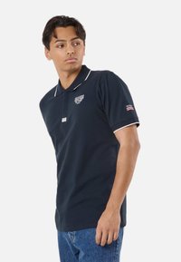 Jeune homme portant un polo bleu marine foncé avec une bordure blanche et du texte, debout avec un bras le long du corps et regardant sur le côté.