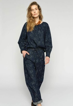 Žena s vlnitými vlasmi, oblečená v tmavomodrom vrchu s kvetinovým vzorom a v rovnakej farbe nohaviciach, s rukou v vrecku a hrubým jantarovým náhrdelníkom.