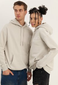 Dva mlada odrasla v bež kapucarjih z žepi spredaj, stojita blizu, eden z rokami v žepih, nevtralno ozadje.