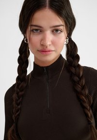 Brun dragkedja tröja med hög krage och ribbad textur. Modellen har långt flätat hår och silverfärgade hoopörhängen.