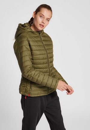 Kobieta ubrana w oliwkowo-zieloną pikowaną kurtkę puchową z kapturem, pozująca z rękami przy rękawach na jednolitym jasnoszarym tle.