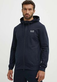 Felpa con zip di colore navy in tessuto morbido, dotata di cappuccio con cordino e logo EA7 sul petto; presenta tasche frontali e polsini a costine.