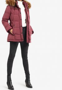 Chaqueta acolchada color marrón oscuro con capucha adornada con pelo, cierre de cremallera y botones, bolsillos laterales y puños acanalados, combinada con vaqueros negros y botines.