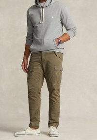 Felpa grigia con cappuccio e coulisse, pantaloni cargo verdi oliva con molte tasche e sneaker bianche su uno sfondo neutro.