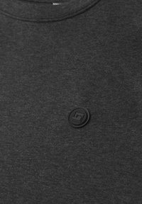 Gros plan sur un tissu gris foncé avec un petit patch circulaire en caoutchouc noir présentant un logo géométrique en relief près du centre.