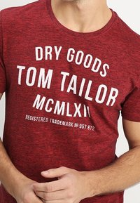 Rode melange T-shirt met een ronde hals. Voorzien van vetgedrukte witte tekst: "DRY GOODS TOM TAILOR MCMLXII" en een geregistreerd handelsmerk eronder. Zachte stofstructuur.