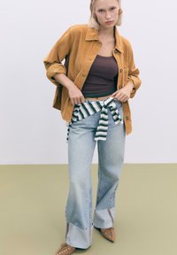 Giacca in corduroy color senape, canotta scura, jeans larghi blu chiaro con tessuto a righe legato in vita e scarpe beige a punta.