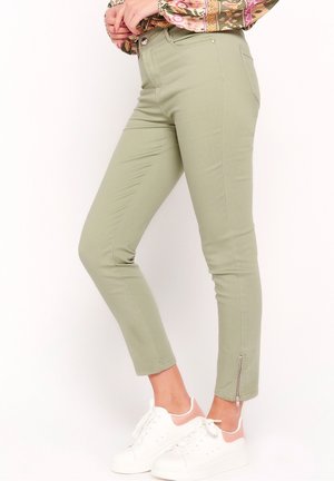 Pantaloni slim fit verde chiaro con cerniera alla caviglia abbinati a sneaker bianche e rosa, indossati da una persona vista di profilo.