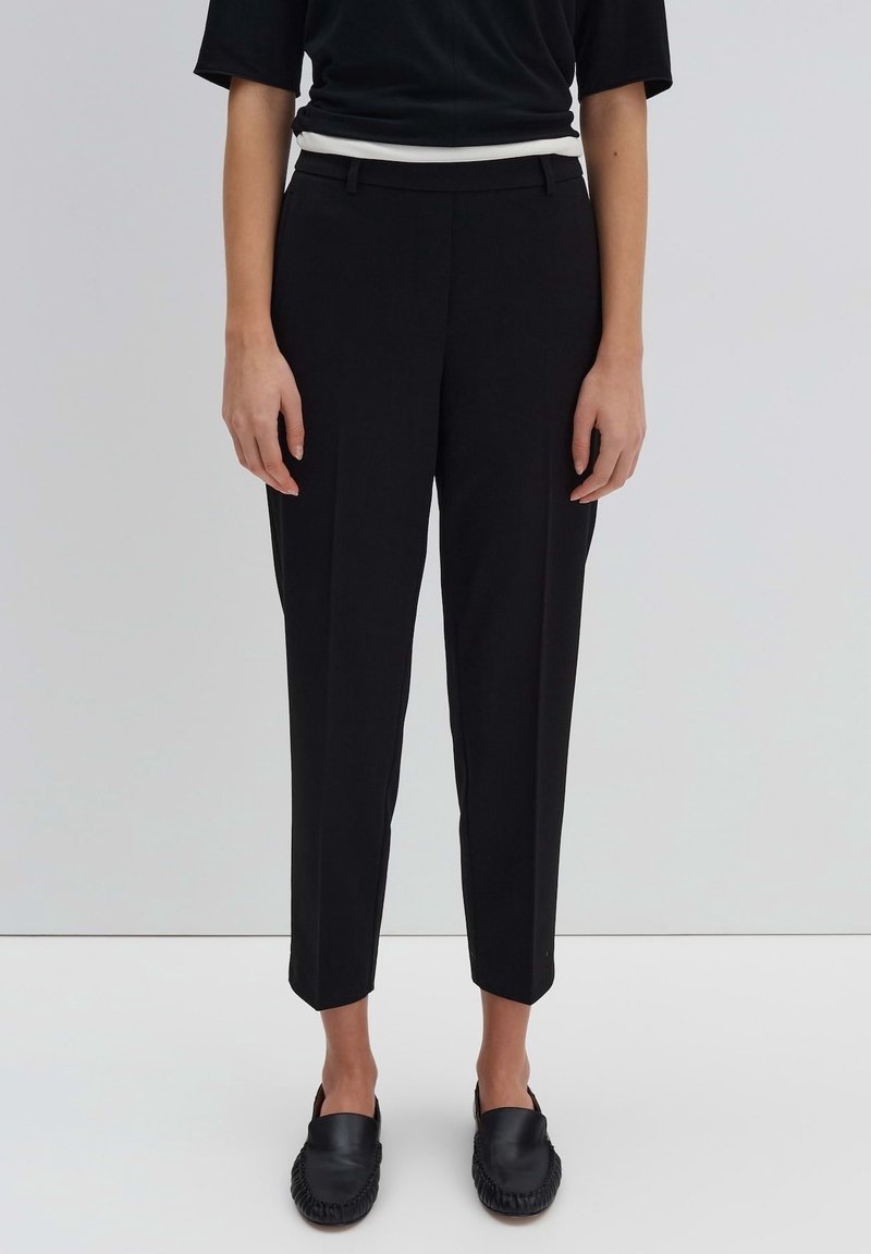 Personne portant un pantalon noir coupé sur mesure, des chaussures noires sans lacets et un haut noir avec un bord blanc à la taille, debout sur un sol gris uni.