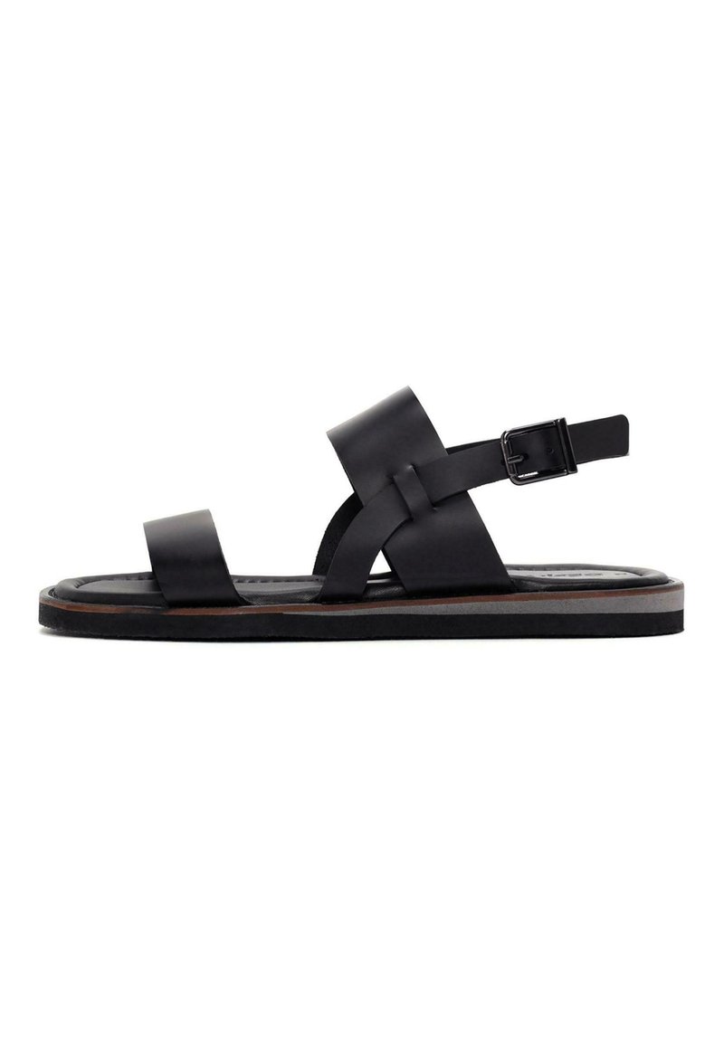 Black leather sandal with wide straps, a buckle closure, and a flat sole featuring a brown accent around the edge. Simple and minimal design.