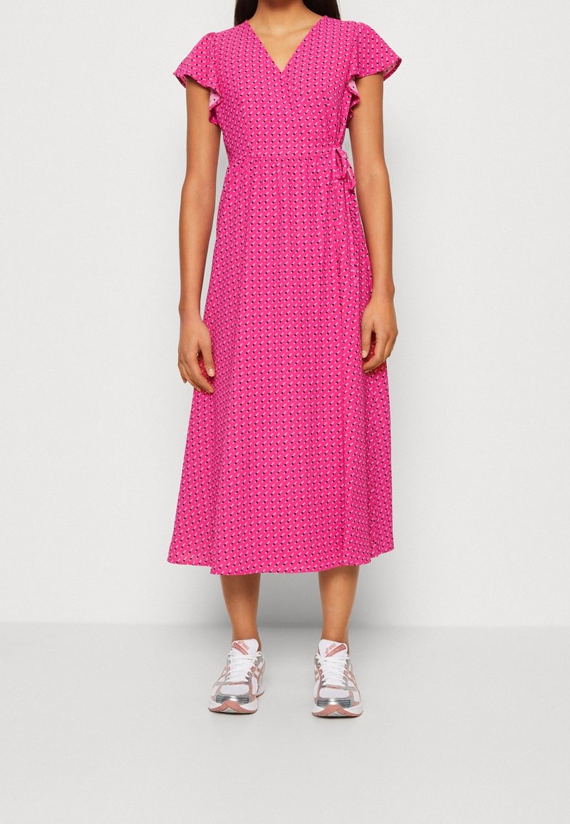 Femme portant une robe portefeuille midi rose avec petits motifs, manches volantes et baskets blanc-rose sur fond uni.