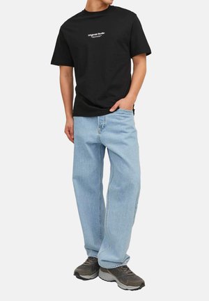 Schwarzes Baumwoll-T-Shirt mit weißem "Original Studio"-Logo, kombiniert mit hellblauen gerade geschnittenen Jeans und dunklen Sportschuhen.