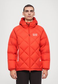 Red puffer jacket with a geometric quilted pattern, hood, two side zippered pockets, and EA7 logo on the left chest. Smooth texture.