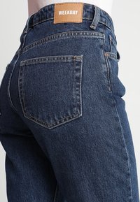 Jeans de mezclilla azul oscuro de cintura alta, con una etiqueta de cuero marrón en la parte posterior que dice "WEEKDAY" y clásicos bolsillos traseros.