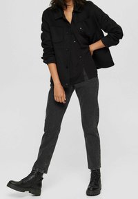 Veste noire avec fermeture à boutons et poches poitrine, superposée à une chemise noire, assortie à un jean gris et des bottes noir à lacets.