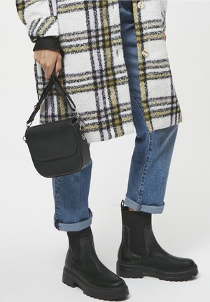 Botas de cuero negras hasta el tobillo con una suela gruesa, combinadas con un bolso cruzado negro texturizado. El conjunto incluye un abrigo de cuadros y jeans de denim remangados.