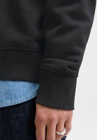 Süvendatud käsi, mis kannab hõbedast sõrmust, toetub küljele; nähtavad on musta dressipluusi varrukas ja sinise teksasärgi varrukas.