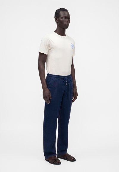 Homme debout portant une chemise blanche à manches courtes avec un petit motif de tortue bleue, un pantalon bleu marine à cordon et des sandales marron sur fond blanc.