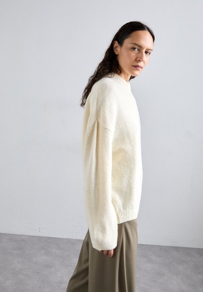 Kremowy, oversize sweter z wysokim kołnierzem i prążkowanymi mankietami. Teksturowany materiał o miękkim wyglądzie, zestawiony z szerokimi szarymi spodniami.